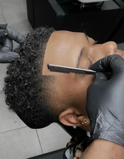 Photo d'un homme dans un salon de coiffure pour homme en Martinique. Le Barber termine les contours de cheveux d'un jeune étudiant aux cheveux texturés avec une lame Gillette.